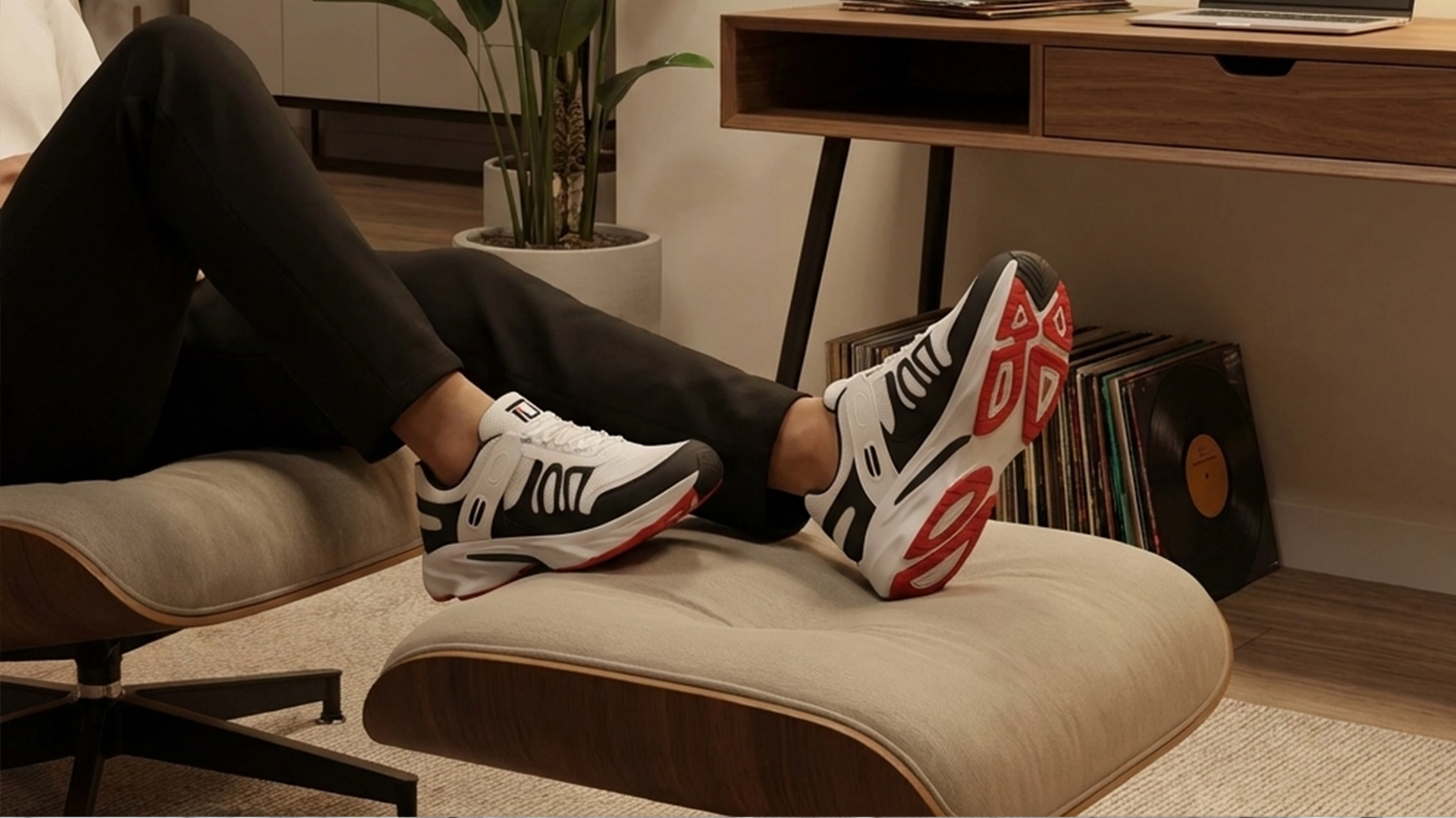 Person wearing black and white sneakers with red accents sitting on a beige ottoman in a home office setting.